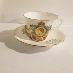 Op de afbeelding: Een wit theekopje en schotel met een gouden rand. Het kopje en de schotel hebben een kleurrijk beeld van koningin Elizabeth II met de tekst "EIIR" en "1952-2002" aan de voorkant.