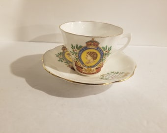 Könnte beinhalten: Eine weiße Teetasse und Untertasse mit goldfarbenem Rand. Tasse und Untertasse zeigen ein farbenfrohes Bild von Königin Elisabeth II mit dem Text "EIIR" und "1952-2002" auf der Vorderseite.