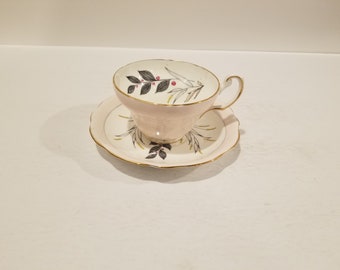 May include: A pink teacup and saucer with a floral design in black, gray, and gold. The floral design features leaves and berries.