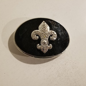 Pode incluir: Uma fivela de cinto prateada com fundo de esmalte preto e um design de flor-de-lis prateada com strass.