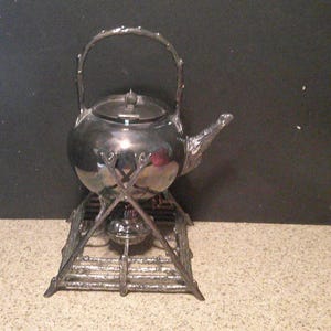 May include: A silver-colored tea kettle with a decorative handle and spout, resting on a matching stand. The kettle has a rounded body and a lid. The stand has a decorative, openwork design. The background is dark.