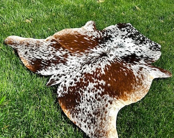 May include: A brown and white cowhide rug with a spotted pattern. The rug is laid out on a green grassy surface.