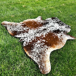 May include: A cowhide rug with a natural pattern of brown, white, and black spots. The rug is irregularly shaped and lies on a bed of green grass, showcasing its unique design and texture.