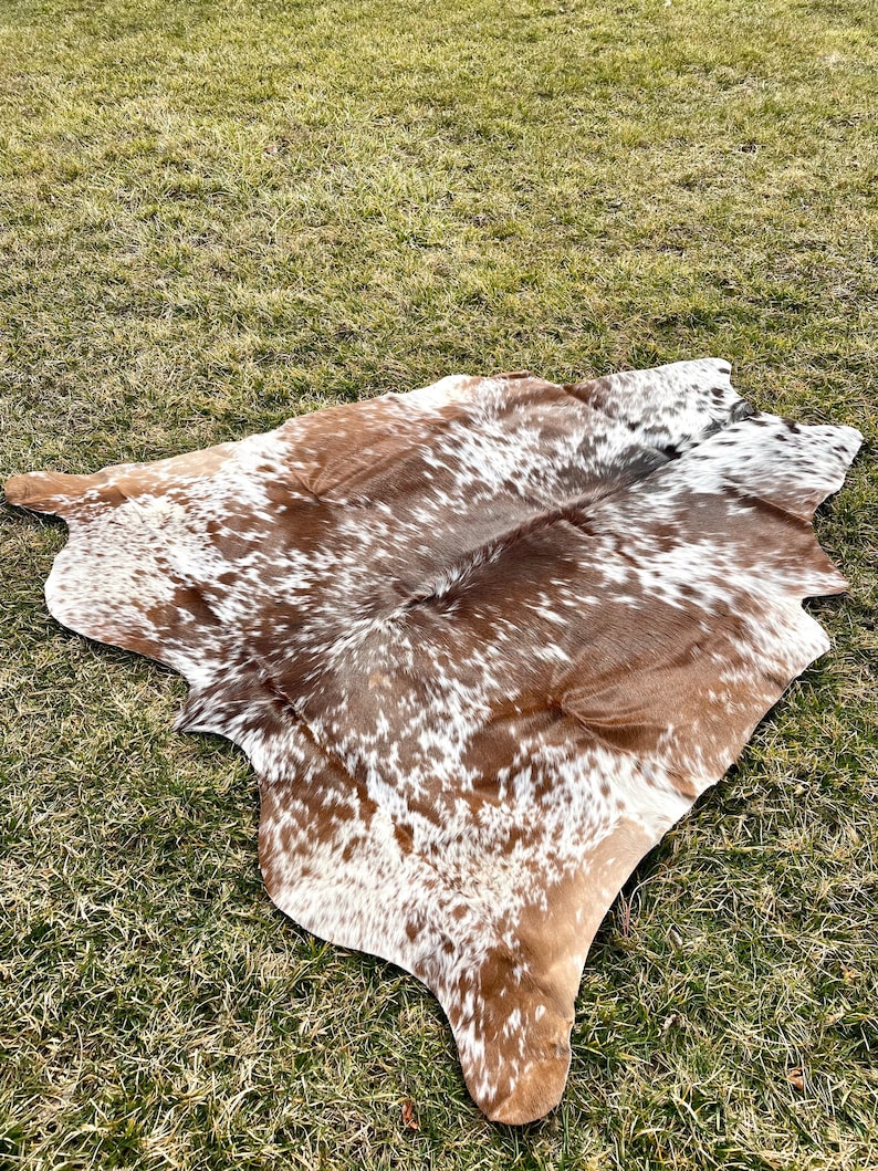 May include: A cowhide rug with a natural pattern of brown and white spots. The rug is irregularly shaped and lies on a bed of green grass, showcasing its organic form and texture. The hide appears soft and supple.