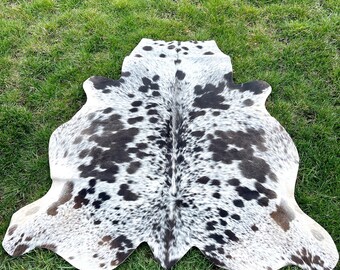May include: A black and white cowhide rug with a spotted pattern. The rug is laid out on a green grassy surface.