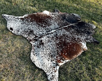 May include: A cowhide rug with a natural pattern of brown and white spots. The rug is irregularly shaped and lies on a bed of green grass, showcasing its unique texture and design. The hide is a natural, rustic home decor item.