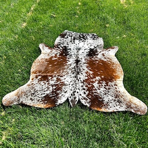 May include: A cowhide rug with a natural pattern of brown, white, and black spots. The rug is irregularly shaped, with rounded edges and a central spine. It is displayed on a bed of green grass.