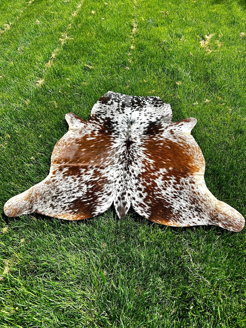 May include: A cowhide rug with a natural pattern of brown, white, and black spots. The rug is irregularly shaped, with rounded edges and a central spine. It is displayed on a bed of green grass.
