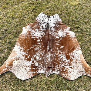 May include: A cowhide rug with a natural pattern of brown, white, and black spots. The rug is irregularly shaped, with a smooth, soft texture. The hide is laid out on a bed of green grass.