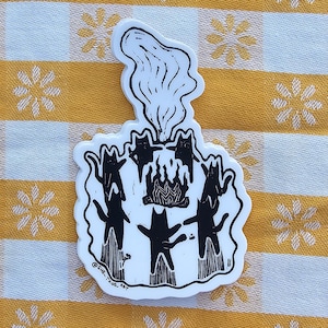Könnte beinhalten: Ein schwarz-weißer Aufkleber mit einer stilisierten Illustration von drei Katzenfiguren, die sich um einen Kuchen mit Rauch versammeln. Der Aufkleber liegt auf einer gelb-weiß karierten Tischdecke.