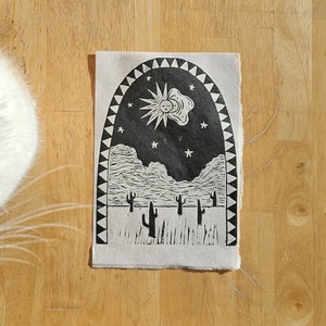Puede incluir: Una impresión de linóleo en blanco y negro de un paisaje desértico con un sol y estrellas en el cielo. La impresión está enmarcada por un borde decorativo.
