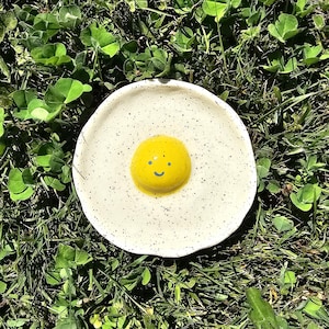 Può includere: Un piatto in ceramica rotondo color bianco sporco con una consistenza maculata, caratterizzato da un centro rialzato giallo brillante e lucido a forma di tuorlo d'uovo. Il tuorlo giallo presenta un piccolo viso sorridente blu dipinto a mano.