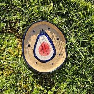 May include: A small, handmade ceramic dish with a beige base and a blue rim. The center features a stylized design with a red and white center, surrounded by a blue outline. Small blue stars are scattered around the rim.