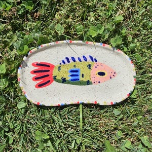 May include: An oval-shaped ceramic tray with a speckled off-white finish. The center features a hand-painted fish in shades of green, blue, pink, and red. The rim of the tray is decorated with a series of small, colorful dots in red, blue, and yellow.