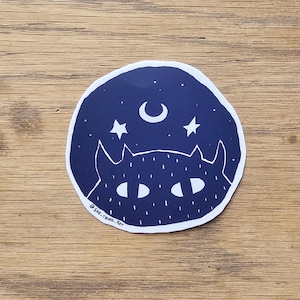 May include: A dark blue sticker with a white outline features a stylized illustration of a cat with its eyes peeking out from behind a crescent moon and stars. The text "@SHE-TRIED.ART" is at the bottom of the sticker.