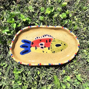 Puede incluir: Un plato ovalado de cerámica con un esmalte beige moteado que presenta un diseño de pez pintado a mano en rojo, amarillo y verde con detalles en azul. El borde está decorado con un patrón repetitivo de pequeños puntos rojos y azules.