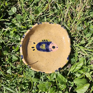 Op de afbeelding: Een rond, gespikkeld beige keramieken schaaltje met een onregelmatige, handgevormde rand. In het midden staat een geschilderde vis met een donkerblauw lichaam, gele vinnen en een lichtroze kop. De rand is versierd met kleine donkerblauwe stippen.