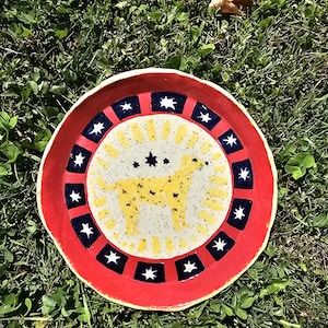May include: A round ceramic plate with a speckled cream center featuring a yellow dog silhouette and a small star. The border is red with a repeating pattern of dark blue squares containing white stars, creating a vibrant and colorful design.