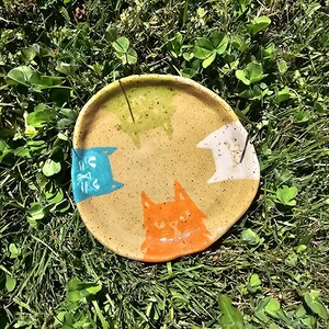 May include: A round, speckled tan ceramic dish featuring four hand-painted cat faces along the rim. The cats are coloured in blue, green, white, and orange, each with simple line-drawn facial features. The dish has a smooth, glossy finish.