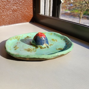 May include: A small, green ceramic dish with a raised, blue and red, volcano-shaped piece in the center.