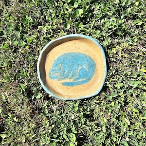 Op de afbeelding: Een ronde, handgevormde keramische schaal met een gespikkelde beige basis en een lichtblauwe rand. Het midden bevat een gestileerde, handgeschilderde blauwe afbeelding van een opgerolde kat. De schaal heeft een onregelmatige rand.
