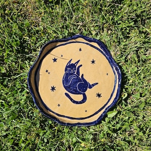 May include: A round, hand-formed ceramic dish with a tan, speckled base and a dark blue, wavy, raised rim. The center features a dark blue illustration of a cat lying on its back, surrounded by small, dark blue star-shaped symbols.