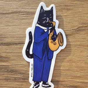 K&ouml;nnte beinhalten: Eine schwarze Katze in einem blauen Anzug spielt Saxophon in Gold. Die Katze hat einen ernsten Gesichtsausdruck. Der Sticker hat den Text "@SHE_TRED_ART" in der linken unteren Ecke.