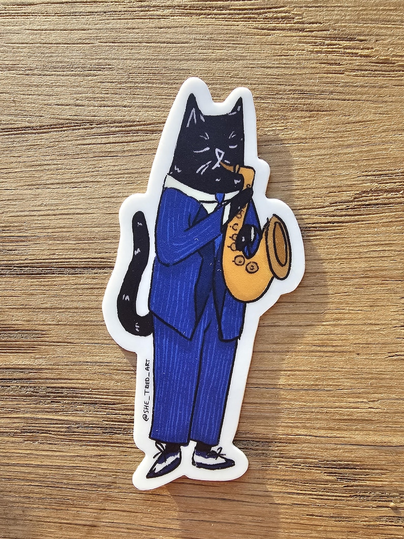 K&ouml;nnte beinhalten: Eine schwarze Katze in einem blauen Anzug spielt Saxophon in Gold. Die Katze hat einen ernsten Gesichtsausdruck. Der Sticker hat den Text "@SHE_TRED_ART" in der linken unteren Ecke.