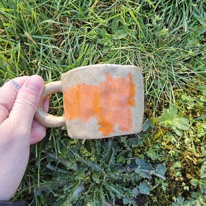 May include: A handmade ceramic mug with a beige base and an orange rectangular design. The mug has a rounded handle and is set against a backdrop of green grass. This unique mug is perfect for enjoying your favorite hot beverages.