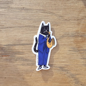 K&ouml;nnte beinhalten: Eine schwarze Katze in einem blauen Anzug mit Krawatte spielt Saxophon in Gold. Die Katze tr&auml;gt schwarze Schuhe. Der Sticker hat den Text "@owl_tree_art" in der rechten unteren Ecke.
