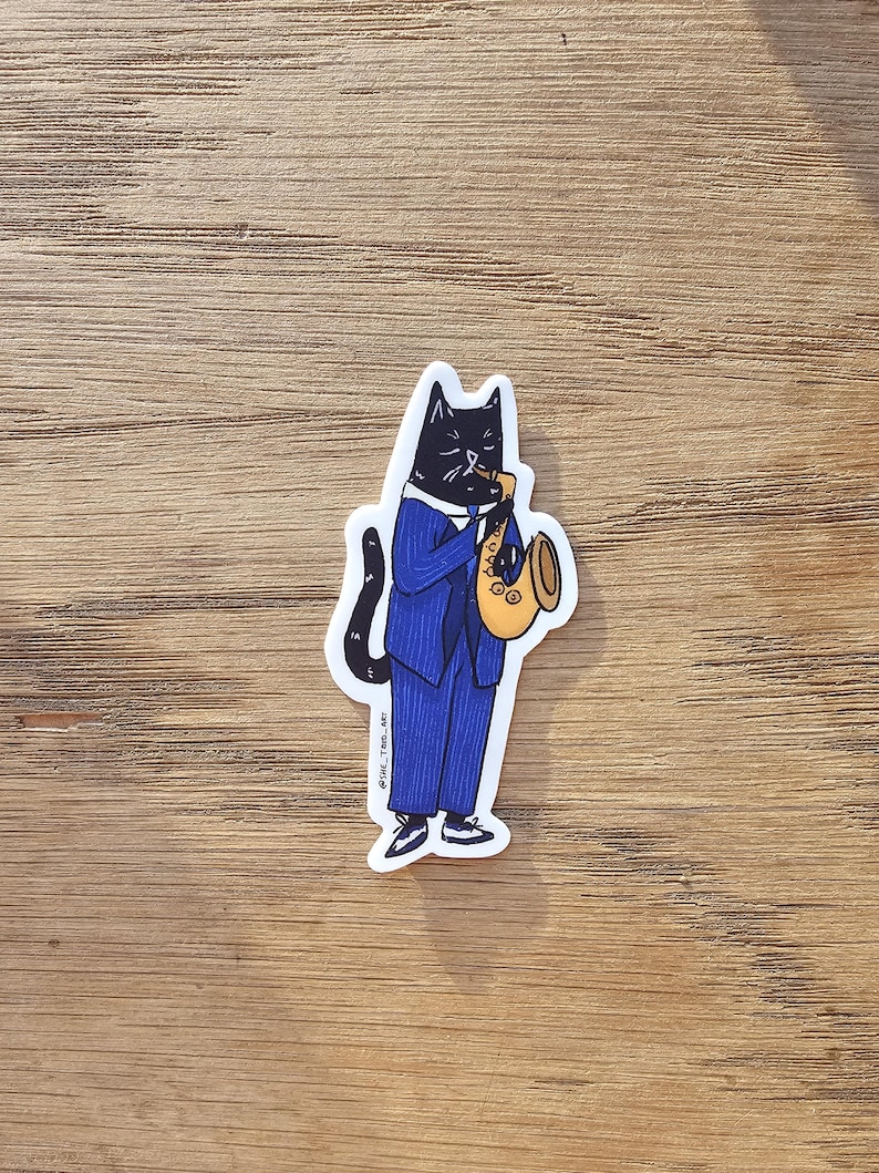 K&ouml;nnte beinhalten: Eine schwarze Katze in einem blauen Anzug mit Krawatte spielt Saxophon in Gold. Die Katze tr&auml;gt schwarze Schuhe. Der Sticker hat den Text "@owl_tree_art" in der rechten unteren Ecke.