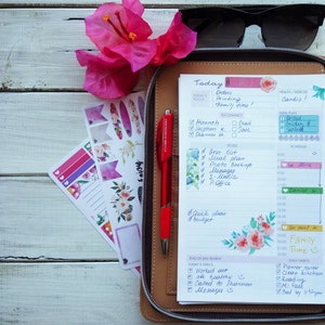 May include: A close-up of an open daily planner with a brown leather cover. The planner is open to the daily page for today, with handwritten to-do lists, contact information, and a meal plan. The planner also includes a section for end-of-day review and daily habits. The planner is decorated with floral stickers and a red pen.