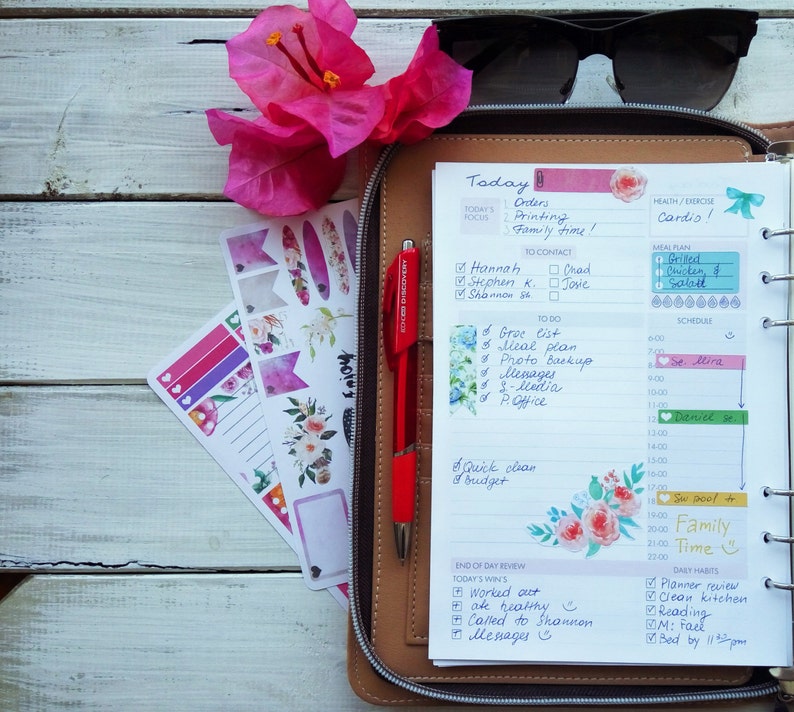 May include: A close-up of an open daily planner with a brown leather cover. The planner is open to the daily page for today, with handwritten to-do lists, contact information, and a meal plan. The planner also includes a section for end-of-day review and daily habits. The planner is decorated with floral stickers and a red pen.