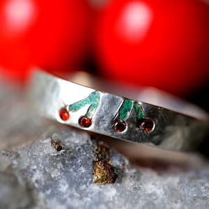 Peut inclure: Une bague en argent avec un motif de cerise. La bague présente trois pierres précieuses rouges représentant des cerises, avec des accents verts pour les feuilles. La bague est posée sur un fond flou avec deux sphères rouges.
