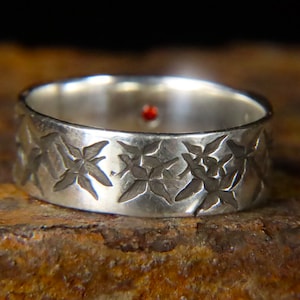 May include: A silver band ring with a textured design of repeating star-shaped patterns. The ring has a small red stone set in the centre of the band.