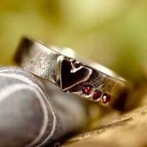 Puede incluir: Un anillo de banda de plata con un acabado texturizado. El anillo presenta un diseño en forma de corazón con un centro negro y cuatro pequeñas piedras preciosas rojas.