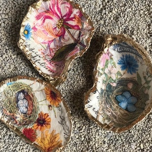 May include: Three decorative oyster shells with gold gilded edges. Each shell features a different floral and botanical design with a decoupage technique. The designs include flowers, nests, and script. The shells are arranged on a bed of small, gray pebbles.