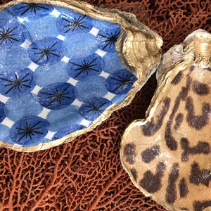 May include: Two decorative oyster shells. One shell features a blue and white pattern with black starbursts. The other shell has a tan and brown leopard print design. Both shells have gold painted edges.