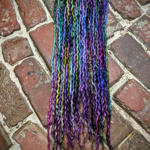 Op de afbeelding: Een set kleurrijke synthetische dreadlocks in tinten paars, blauw, groen en zwart. De dreadlocks zijn in een rechte lijn gerangschikt en liggen op een bakstenen oppervlak.