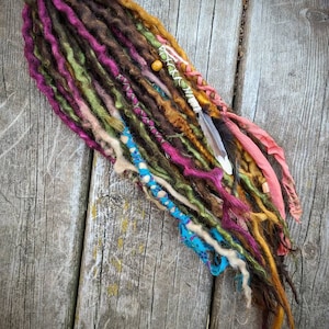 Puede incluir: Un conjunto de rastas sintéticas de colores con cuentas, una pluma y detalles de tela. Las rastas están dispuestas sobre una superficie de madera.
