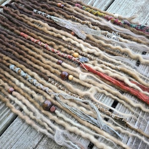 May include: A collection of brown, blonde, and white synthetic dreadlocks with beads and fabric accents. The dreadlocks are arranged in rows on a wooden surface.