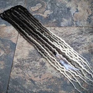 May include: A set of long, synthetic dreadlocks with a black to white ombre colour gradient. The dreadlocks are arranged in a straight line on a textured grey surface. A small white feather is attached to one of the dreadlocks.