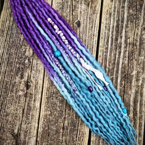 May include: A set of long, ombre synthetic dreadlocks in shades of purple and blue. The dreadlocks are decorated with white string and turquoise beads.