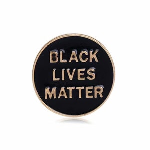 May include: A round, black enamel pin with gold-colored text that reads "BLACK LIVES MATTER." The pin has a gold-colored border and is set against a white background. This pin is a statement piece.