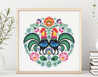 May include: A colorful folk art design featuring two roosters with green, blue, red, yellow, and black accents. The roosters are surrounded by a circle of flowers and leaves.