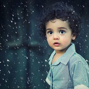 Peut inclure: Un jeune enfant aux cheveux bouclés foncés portant une chemise boutonnée bleu clair regarde directement la caméra. L'arrière-plan est bleu foncé avec des gouttes de pluie blanches qui tombent.