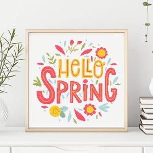 Puede incluir: Una obra de arte en punto de cruz enmarcada con las palabras "HELLO SPRING" en amarillo y rosa. El texto está rodeado de diseños florales y de hojas en rosa, amarillo, verde y azul. La obra se exhibe sobre una superficie blanca.