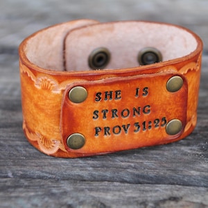 May include: A brown leather cuff bracelet with a metal plate engraved with the text "SHE IS STRONG PROV 31:25".
