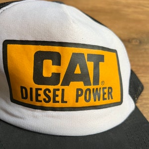 May include: White and black trucker hat with a yellow rectangle featuring the black text "CAT DIESEL POWER". The hat has a white front panel, a black brim, and a black mesh back.