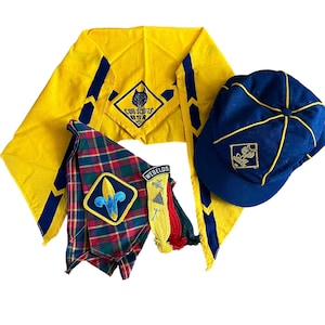May include: A collection of Cub Scout BSA items, including a yellow neckerchief with blue accents and a wolf emblem, a blue cap with a gold emblem, and two plaid neckerchiefs with patches. Also included are several merit badges.
